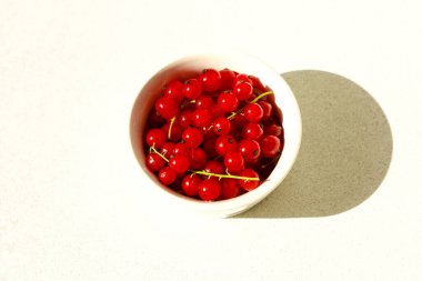 Bir kase birçok küçük kırmızı frenk üzümü barındırır. Kase beyazdır ve hafif bir yüzeye yerleştirilmiştir. Hava güneşli, renkler arka planda göze çarpıyor..