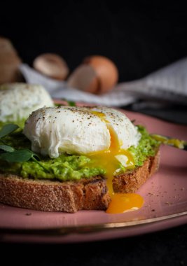 Kahvaltı: haşlanmış yumurta, yeşil avokado kremalı bıçakla pembe tabakta kızarmış ekmek. Arka planda yumurta, ekmek ve havlu var. Tabakta sıvı yumurta sarısı.