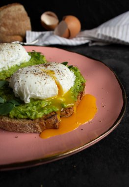 Taze avokado kremalı ekmek üzerine iki haşlanmış yumurta. Sıvı yumurta sarısı pembe tabakta yumurtadan çıkar. Arka planda yumurta, ekmek ve havlu var..