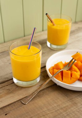 İki bardakta taze mango lassi, metal pipetler ve ahşap kırsal masada meyveli mango meyvesi.
