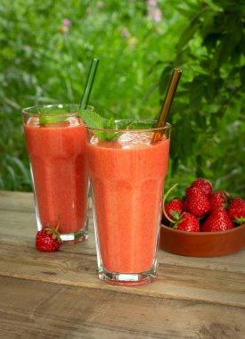 İki bardak çilekli meyve püresi, nane yaprakları ve cam pipetlerle yaz bahçesindeki ahşap bir masada.