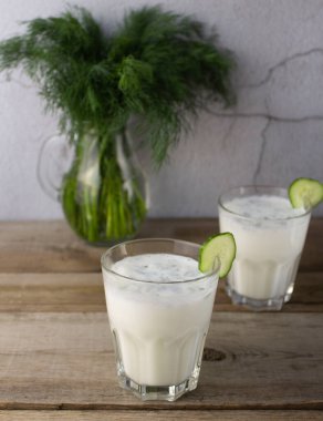 Ayranlı ve dereotlu bir bardak, ahşap bir kırsal masada bir dilim salatalık. Arka planda ayran olan ikinci bardak, vajinanın yanında dereotu olan cam sürahi.