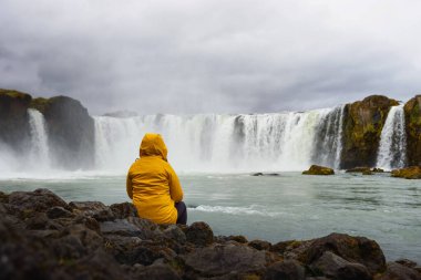Sarı ceketli turist İzlanda 'daki Godafoss şelalesinde dinleniyor.
