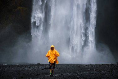 Sarı yağmurluk giyen turist İzlanda 'daki Skogafoss şelalesinden yürüyerek geliyor.