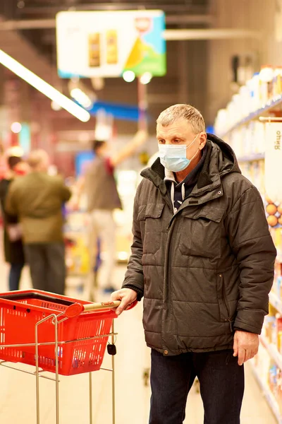 A 65-year-old man in a blue medical mask picks up groceries at a supermarket. Buying food during the coronavirus pandemic.