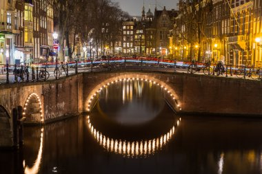 Alacakaranlıkta Amsterdam 'daki Leidsegracht ve Keizersgracht kanallarının kesiştiği köprülerin manzarası. Bisikletler ve binalar görülebilir. Trafiğin izleri bir köprüde görülebilir..
