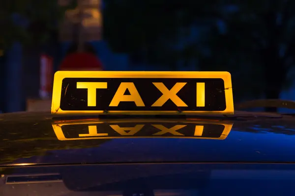 Closeup to a yellow and black taxi sign on a top of a car. Reflections can be seen on the car roof.