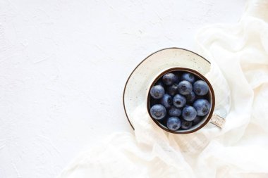 Bir fincanda çiğ yaban mersini ve çay tabağı ve beyaz masa örtüsü ve doku arka planı, düz yatak, kopyalama alanı. Sağlıklı yeme kavramı.