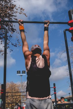 Formda ve yakışıklı sporcu yazın güneşli bir gününde bir sokak idman parkında ağırlık yeleği ile spor yapıyor (barfiks çekiyor).