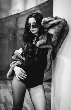 Siyah tulumlu şık esmer model, çorap, eko kürk manto, güneş gözlüğü ve bakır ve taş duvarlarda poz veren topuklu ayakkabı (siyah ve beyaz).)