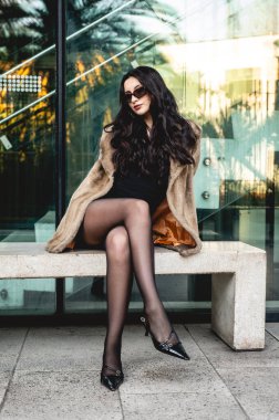 Siyah tulumlu şık esmer manken, güneş gözlüğü, güneş gözlüğü, eko kürk ve topuklu ayakkabılar güneşli meydanda taş bankta poz veriyor.