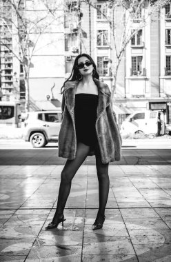 Siyah tulumlu şık esmer model, çorap, güneş gözlüğü, eko kürk manto ve topuklu ayakkabılar bir sokak önünde geçen araçlar (siyah beyaz))