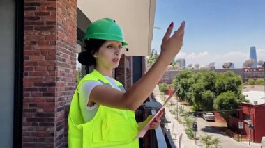 Güvenli yeşil kask ve yansıtıcı yelek giyen profesyonel bir kadın inşaat alanındaki balkondan talimat veriyor.