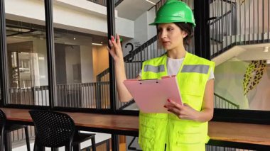 Güvenli yeşil kask ve yansıtıcı yelek giyen profesyonel bir kadın şantiyedeki ofisinde yönünü mutlu bir şekilde tarif ediyor.