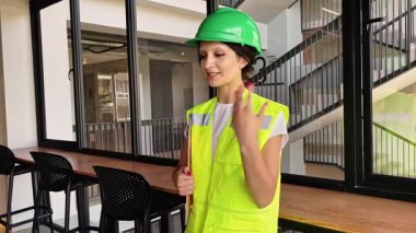 Güvenli yeşil kask ve yansıtıcı yelek giyen profesyonel bir kadın şantiyedeki ofisinde yönünü mutlu bir şekilde tarif ediyor.