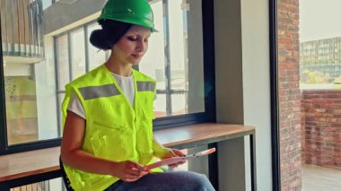 Güvenli yeşil kask ve yansıtıcı yelek giyen profesyonel bir kadın şantiyedeki ofisinde yönünü mutlu bir şekilde tarif ediyor.