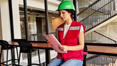 Female construction supervisor wearing safety helmet and vest giving instructions and directing workers at building site holding project folder