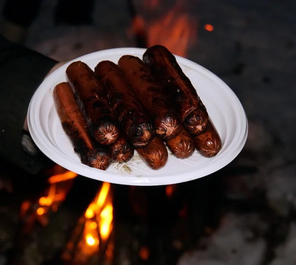 Smoked sausages on white plastic plate on the background of fire — Stock Photo, Image