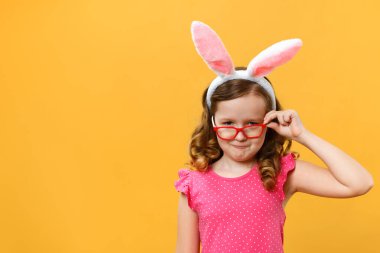 Mutlu Paskalyalar. Tavşan kulaklı şirin bir kız ve sarı arka planda gözlüklü. Çocuk gözlüklerin altından dışarı bakar..