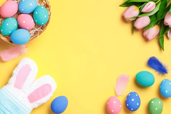 ハッピー イースター バスケットの多色の卵 保護マスクのウサギの耳 黄色の背景にチューリップの花束 コピースペース付きフラットレイグリーティングカード — ストック写真