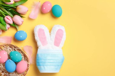 Paskalya konsepti. Koruyucu tıbbi maskeli bir tavşan ve sarı arka planda renkli yumurtalarla dolu bir sepet..