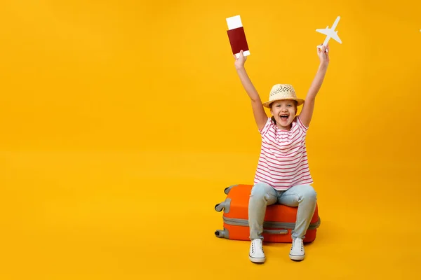 Happy little girl sitting on a suitcase, raised her hands with a paper airplane and a vaccination passport on a yellow background. The child dreams of travel and vacation. Copy space.