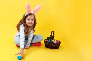 Stüdyoda sarı arka planda tavşan kulaklı küçük bir kız. Tatlı bir çocuk paskalya sepetiyle yerde oturuyor ve yumurta avlıyor..
