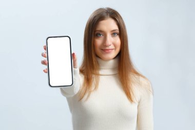 Gülümseyen genç kadın beyaz dokunmatik ekranlı akıllı telefon gösteriyor. Gri arka planda güzel kahverengi saçlı kız..