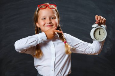 Okula dönelim. Neşeli liseli kız işaret parmağıyla çalar saati gösteriyor. Siyah karatahta arkaplanındaki çocuk.