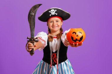 Cadılar Bayramı 'nı kutluyoruz. Stüdyoda mor arka planda bir korsanın karnaval kostümlü neşeli kızı. Çocuğun elinde bir kılıç ve balkabağı kovası var..