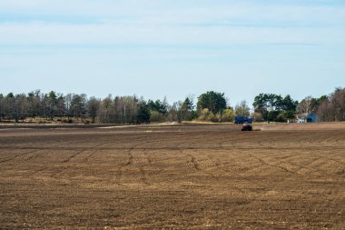Orman kenarında traktör ve mavi römorklu geniş açık tarım arazisi.