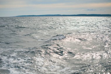 Işıldayan güneş ışığıyla bulutlu gökyüzünün altında uzak kıyı şeridi ve nazik dalgalar.