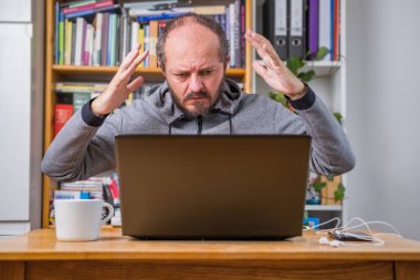 Angry man working online from home office on computer laptop behind vintage desk, arms gesture, business problem concept clipart