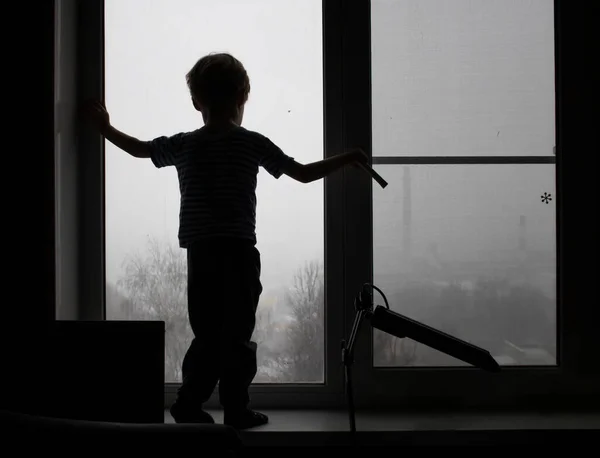 Je to silueta dítěte. Dítě zírající z okna, je nebezpečné pro malé děti zůstat v okně. Zimní deprese, špatné počasí a koncepce nemoci — Stock fotografie