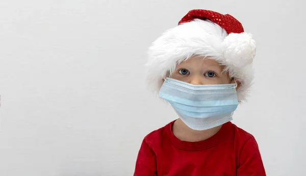 Portrait Un enfant, un tout-petit dans un chapeau rouge de père Noël est malade à Noël. Un garçon portant un masque médical n'ira pas à une matinée à la maternelle à cause de l'épidémie de COVID. L'enfant est triste. Bannière d'espace de copie — Photo