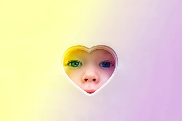 The baby face, eyes and nose peep out of a heart-shaped hole in the center. A child looks into the camera through a heart hole. Love and health concept, mock up, poster, — Stock Photo, Image