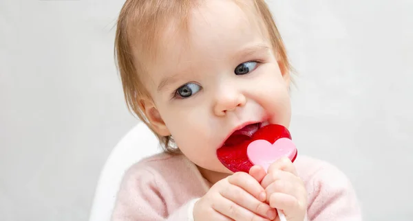 Retrato de un bebé pequeño con caramelos de piruleta rosa en un espacio de copia de pancarta de fondo blanco. Un niño lame un caramelo en forma de corazón. Daño a los dientes —  Fotos de Stock