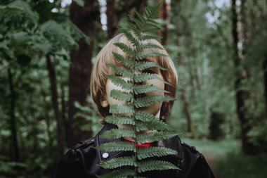 Karanlık bir ormanda bir çocuk eğrelti otu yaprağıyla kaplıdır. Siyah deri ceketli bir çocuk ormanda modaya uygun bir moda derin mat gölgesinde