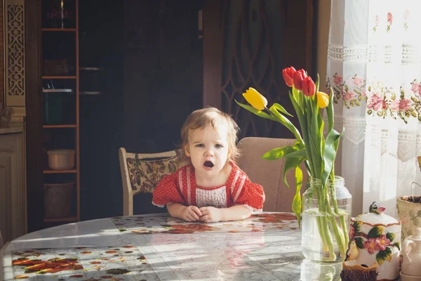 Enojada niña inteligente sentada en una mesa en una cocina campestre con flores festivas y esperando comida — Foto de Stock