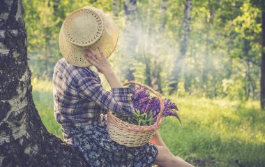 Sırtı dönük bir kız, ormanın yanında duruyor, beyaz giysiler ve hasır şapkalı, elinde bir sepet ekmek.