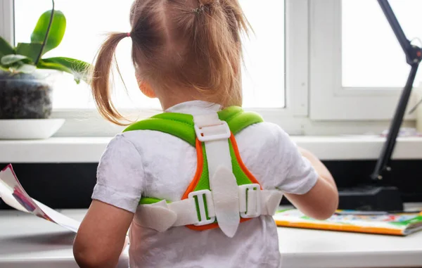 Uma criança uma pequena menina pré-escolar senta-se a uma mesa em um espartilho corretivo para corrigir a postura da coluna vertebral e formar uma coluna correta. Fotografia De Stock