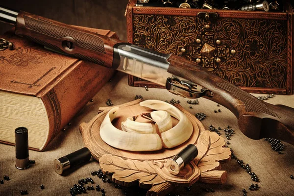 Yine de hayat avlamak: ahşap bir stantta yaban domuzu ganimet dişleri, deri kaplı bir kitap üzerinde av tüfeği, kartuşlarla dekoratif kasa, kaba keten masa örtüsüne kurşun sıkma
