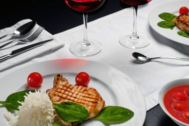 Pirinç garnitürlü beyaz bir tabakta servis edilen domuz pirzolası, iki bardak kırmızı şarap, siyah bir masada beyaz masa örtüsü.