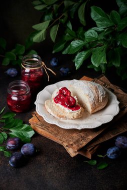 Krem peynirli tostun üstüne renkli erik reçeli. Erik dalları ve eriklerle çevrili, retro tarzı bir gazeteyle eski bir tabakta taze pişmiş ekmek. Masadaki cam kavanozlar.