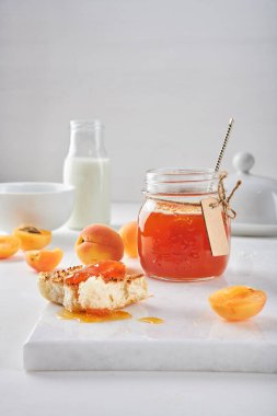 Şirin bir cam kavanozda el yapımı kayısı reçeli ve el işi etiketi. Masada taze çiğ kayısılar var. Kahvaltı ekmeğine biraz reçel bulaşmış. Arka planda bir şişe süt, tereyağı tabağı, seramik kase.