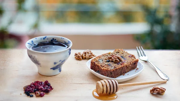 A slice of vegan cake with super fruit and nuts, honey and a cup of tea on wooden table