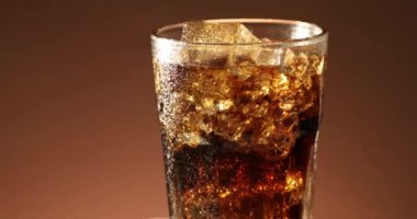 Buzlu kola bardağı yavaşça yakın çekimde dönüyor. Kahverengi arka planda dönen soğuk soda içeceği. 4K video görüntüleri