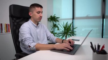 4K, ciddi genç iş adamı ofis masasında modern bir dizüstü bilgisayarla oturuyor ve çalışıyor. Erkek girişimci iş yerindeki bilgisayarda yazıyor. Kafkasyalı adam defter kullanıyor. Masadaki akıllı telefon..