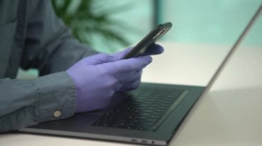 4K, yaratıcı adam eldiven takıyor ve ofisteki bilgisayar klavyesinde akıllı telefonu daktilo ediyor. İş adamı Coronavirus salgını sırasında iş için telefon ve dizüstü bilgisayar kullanıyor. İş büyümesi kavramı.