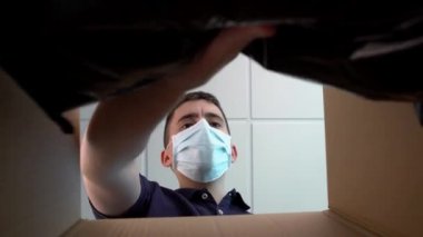 Tıbbi maskeli beyaz bir adamın içeriden görüntüsü çevrimiçi dükkandan sipariş edildi. İşçi siyah paketli bir karton kutu hazırlıyor. İzolasyon karantina koronavirüs kilitleme kavramı.